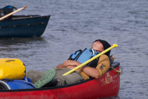 Lucien rests near the shore in his canoe, lying back with his eyes closed.