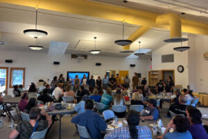 A large, indoor gathering in Mayo with community seated at tables, listening to trip participants who stand in front a photo of the group on the Wind.