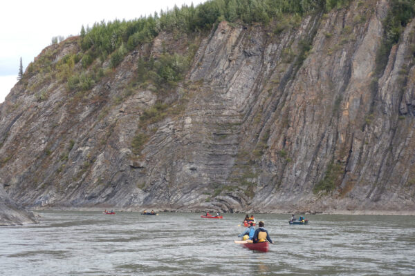 Youth paddle down the Wind River, passing tall, layered cliffs that rise dramatically from the banks of the Peel River.