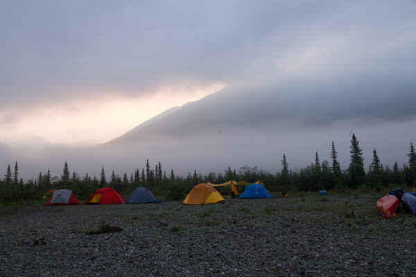 Smoke hides the mountains behind tents scattered along the gravel beach near the river.