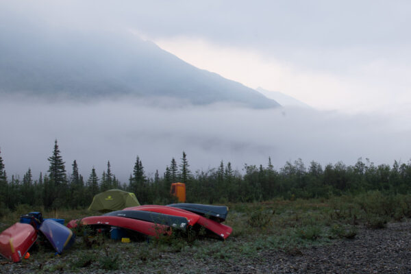 Canoes flipped over near the forest edge, with low clouds and smoke hanging in the background.