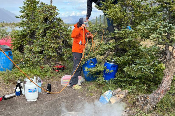 Joti and Ashwin hang up a rope at a campsite while smoke rises from a pot where dinner is being cooked.