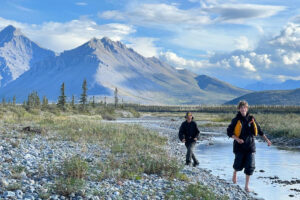 Chayce (left) and Dominic (right) walk along the river bed, with dramatic peaks and clouds in the background.