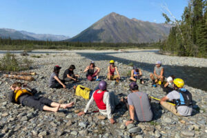 Trip participants sit and chat in a circle on a gravel bar, with the river and mountains behind them. From the front moving clockwise: Laurent (red PFD), Dominic, Kadrienne, Chayce, Joti, Virginia, Chantel, Joel, Bethany, Lucien, and Jona.