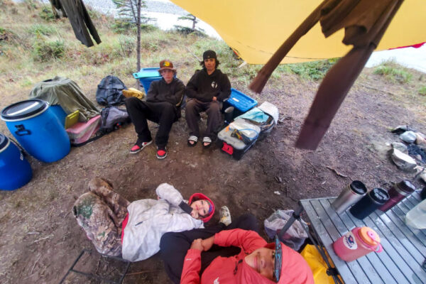 Youth relax under tarps where clothes are hung to dry. Dominic (left) and Chayce (right) sit on chairs in the back, while Lucien lounges on a chair and Shalaya sits on the ground in the front.