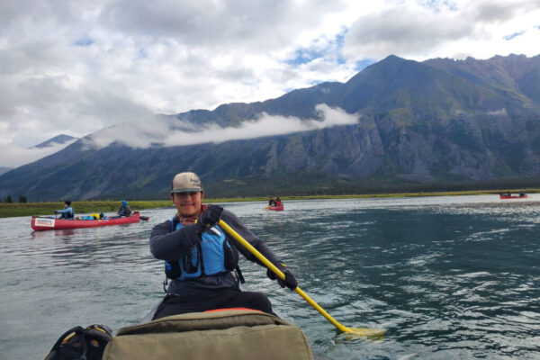 Lucien smiles as he paddles towards the camera, with other participants in canoes scattered behind him.