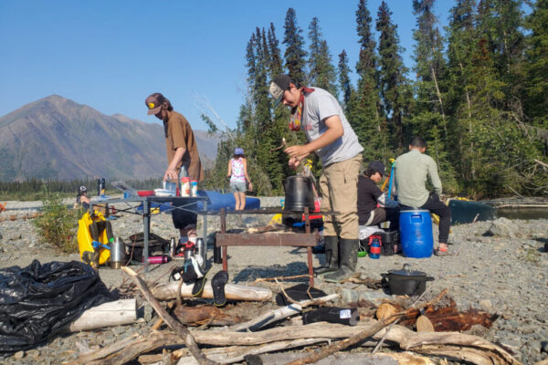 Bethany, Dominic, Shalaya, Lucien, Chayce, and Laurent (from left to right) prepare food and organize gear at a campsite on a sunny day.