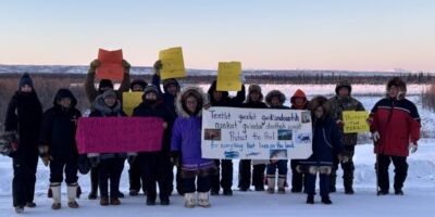 People hold signs advocating for the Peel in Fort McPherson, NWT at sunset, including two large ones that read “This is our land, this is our water, and that’s where we were raised from” and “Protect the Peel for everything that lives on the land.” Teetl'it Gwich'in traditional territory, photo by Laura Nerysoo.