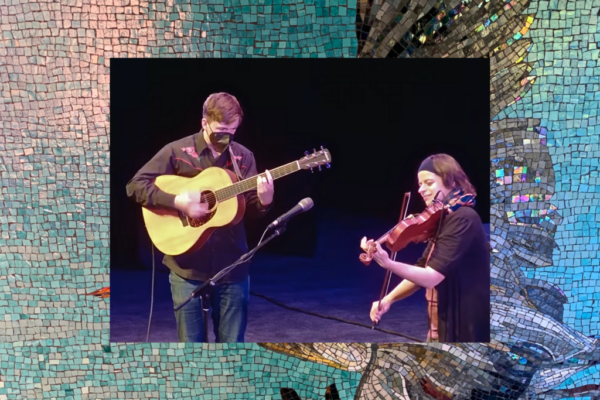 Graeme Poile and Keitha Clark perform on stage, Graeme strums an acoustic guitar and Keitha smiles while playing a fiddle.