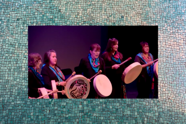 Members of Remmi Elin drum and sing on stage, each holding hand drums and wearing matching blue patterned scarves.