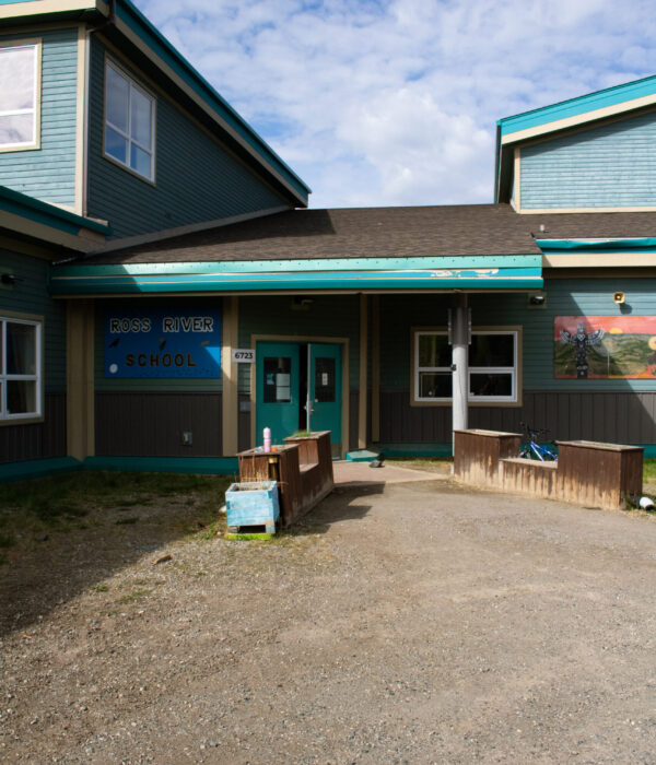 The Ross River blue school building with teal and gray siding, and a mural on the right sidewall. Photo by Hannah Hofsink, Ross River Dena Council territory.