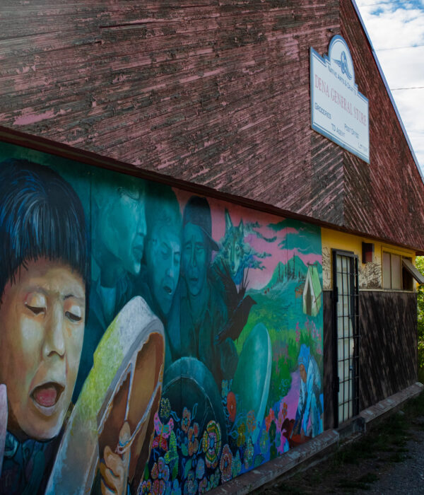 Kaska Dena general store building featuring a colourful mural with blues, greens, and pinks that depicts historical and cultural scenes of people drumming, nature, and symbolic elements. Photo by Hannah Hofsink, Ross River Dena Council territory.