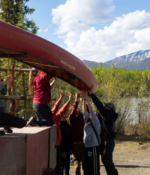 A group of students lift a red canoe off a trailer with trees and mountains in the background. Photo by Hannah Hofsink, Ross River Dena Council territory.