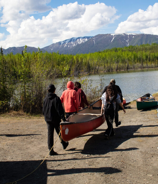 Students carry a red canoe towards the shore of Jackfish Lake, surrounded by green trees and distant snow-capped mountains. Photo by Hannah Hofsink, Ross River Dena Council territory.