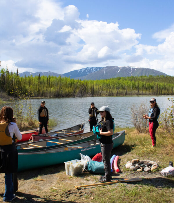 A group of five people prepare for canoeing by the shore of Jackfish Lake with canoes and gear. Photo by Hannah Hofsink, Ross River Dena Council territory.