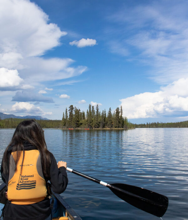A student in a yellow life jacket paddles a canoe on a calm Jackfish Lake towards a small island with tall pine trees. Photo by Hannah Hofsink, Ross River Dena Council territory.