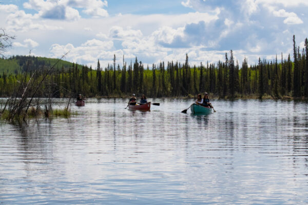 A group of people canoe towards the camera on Jackfish lake, with blue skies reflecting on the calm water. Photo by Hannah Hofsink, Ross River Dena Council territory.
