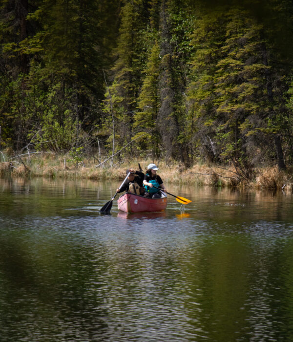 Kadrienne, in the stern, paddles away from a dense, evergreen forest with a Ross River School student in a red canoe. Photo by Hannah Hofsink, Ross River Dena Council territory.