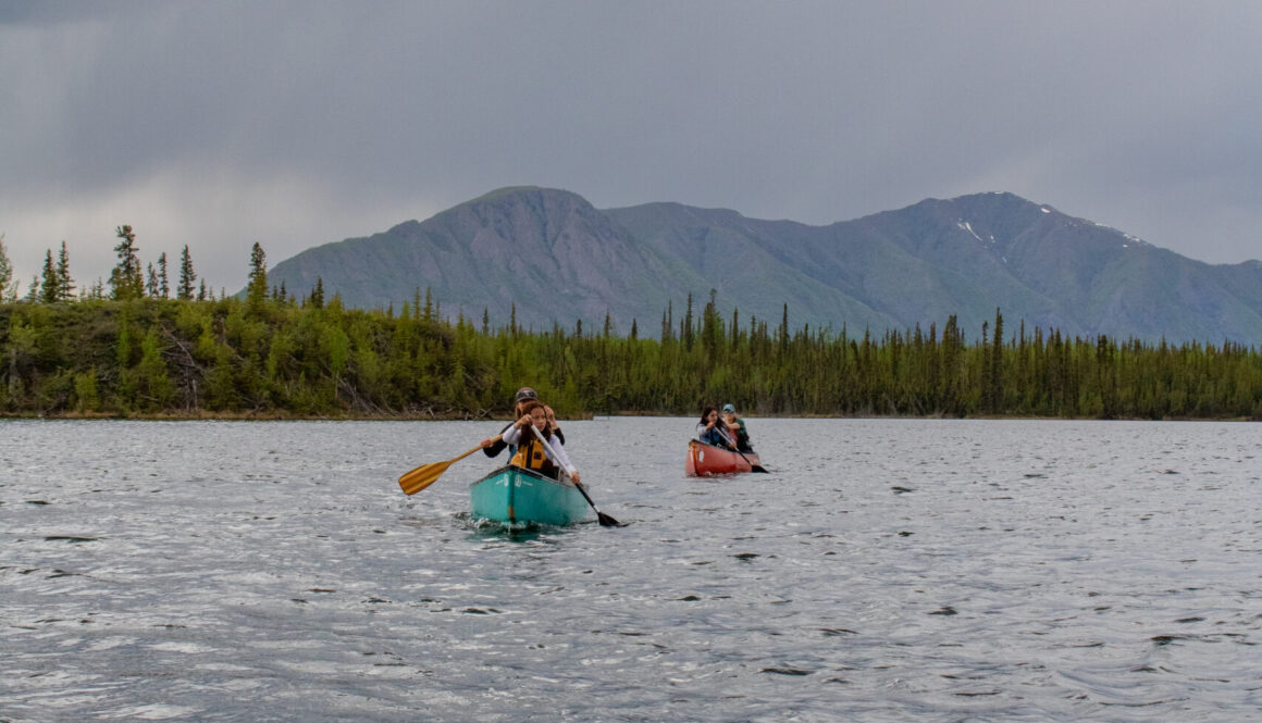 Four people paddle in two canoes on Jackfish Lake with a forested shoreline and mountain in the background on a cloudy day. Photo by Hannah Hofsink, Ross River Dena Council territory.