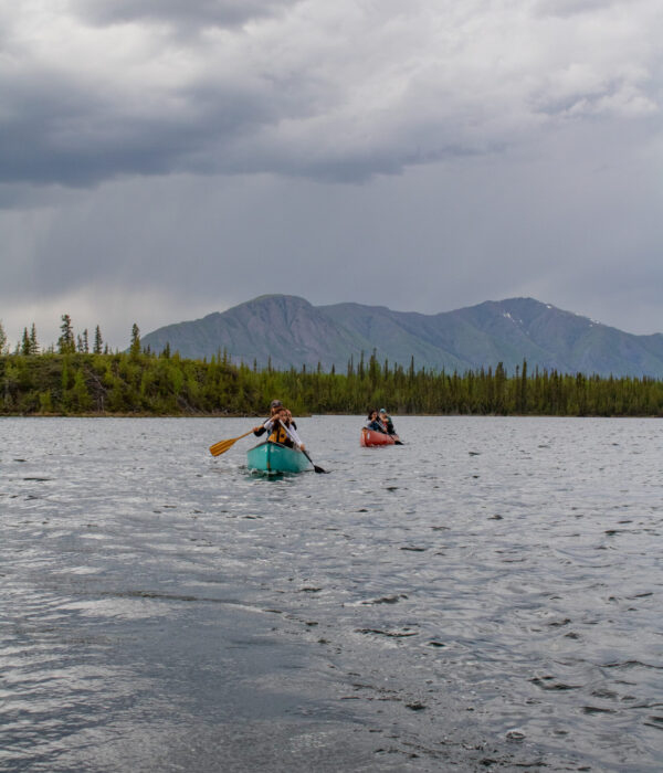 Four people paddle in two canoes on Jackfish Lake with a forested shoreline and mountain in the background on a cloudy day. Photo by Hannah Hofsink, Ross River Dena Council territory.