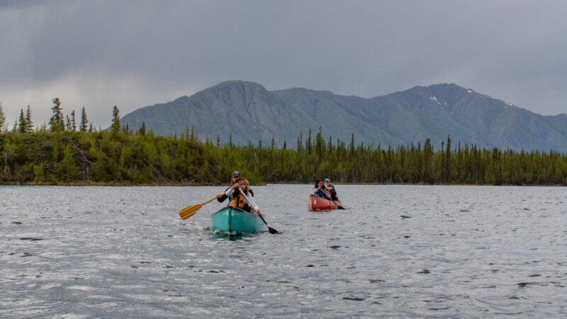 Four people paddle in two canoes on Jackfish Lake with a forested shoreline and mountain in the background on a cloudy day. Photo by Hannah Hofsink, Ross River Dena Council territory.