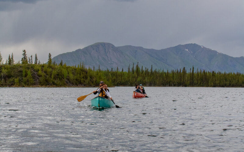 Four people paddle in two canoes on Jackfish Lake with a forested shoreline and mountain in the background on a cloudy day. Photo by Hannah Hofsink, Ross River Dena Council territory.