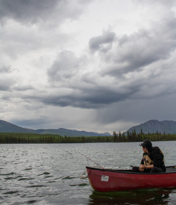A Ross River School student fishes from a red canoe on Jackfish Lake under a cloudy, overcast sky. Photo by Hannah Hofsink, Ross River Dena Council territory.