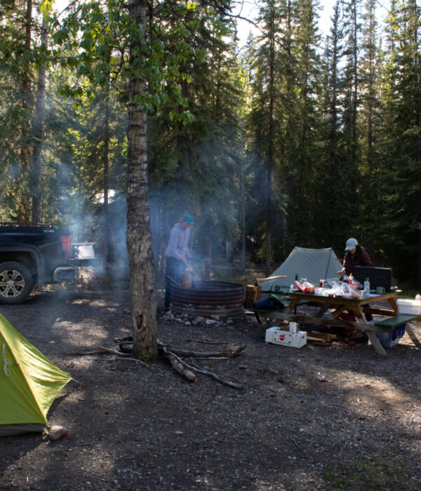 Smoke from a campfire rises beside a green and orange tent while two people make lunch at a picnic table. Photo by Hannah Hofsink, Ross River Dena Council territory.