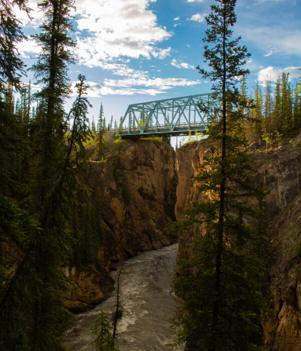 A steel blue truss bridge spans a deep rocky canyon with the Lapie River flowing below. Photo by Hannah Hofsink, Ross River Dena Council territory.